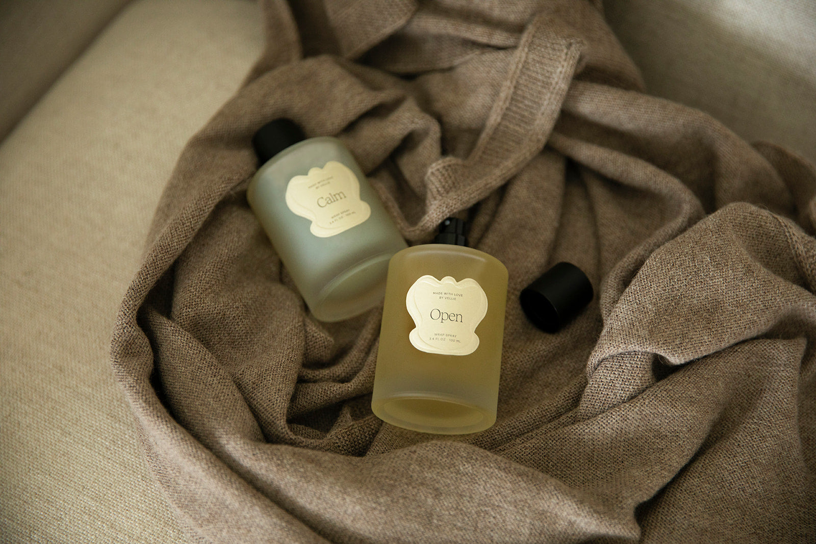 Two wrap sprays scents Open and Calm delicately placed on top of a Vellie Wrap. Created to spray on your wrap. The image is a fragrant reminder of love, making each wrap feel like a personal sanctuary.