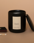 Vellie Open Candle in black vessel with lid off, accompanied by a black Vellie matchbox. This vegan candle offers a bright, balancing fragrance of lemongrass, mate, and thyme.
