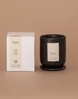 Vellie Open Candle. A luxury candle with herbal, citrus and green notes. Seen here, the chic black vessel next to its packaged box. Box colors are neutral, cream, black and white.