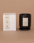 Vellie Hope Candle. A luxury candle with lush, bright and fresh notes. Seen here, the chic black vessel next to it's packaged box. Box colors are neutral, cream, black and white.