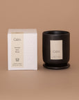 Vellie Calm Candle. A luxury candle with aromatic, spicy and woody notes. Seen here, the chic black vessel next to it's packaged box. Box colors are neutral, cream, black and white.