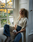 Model holds a cup of tea as she sits inside a window nook looking outside the window. She wears a long ivory wrap that comforts her. Crafted from 100% fair trade, sustainably sourced baby alpaca.