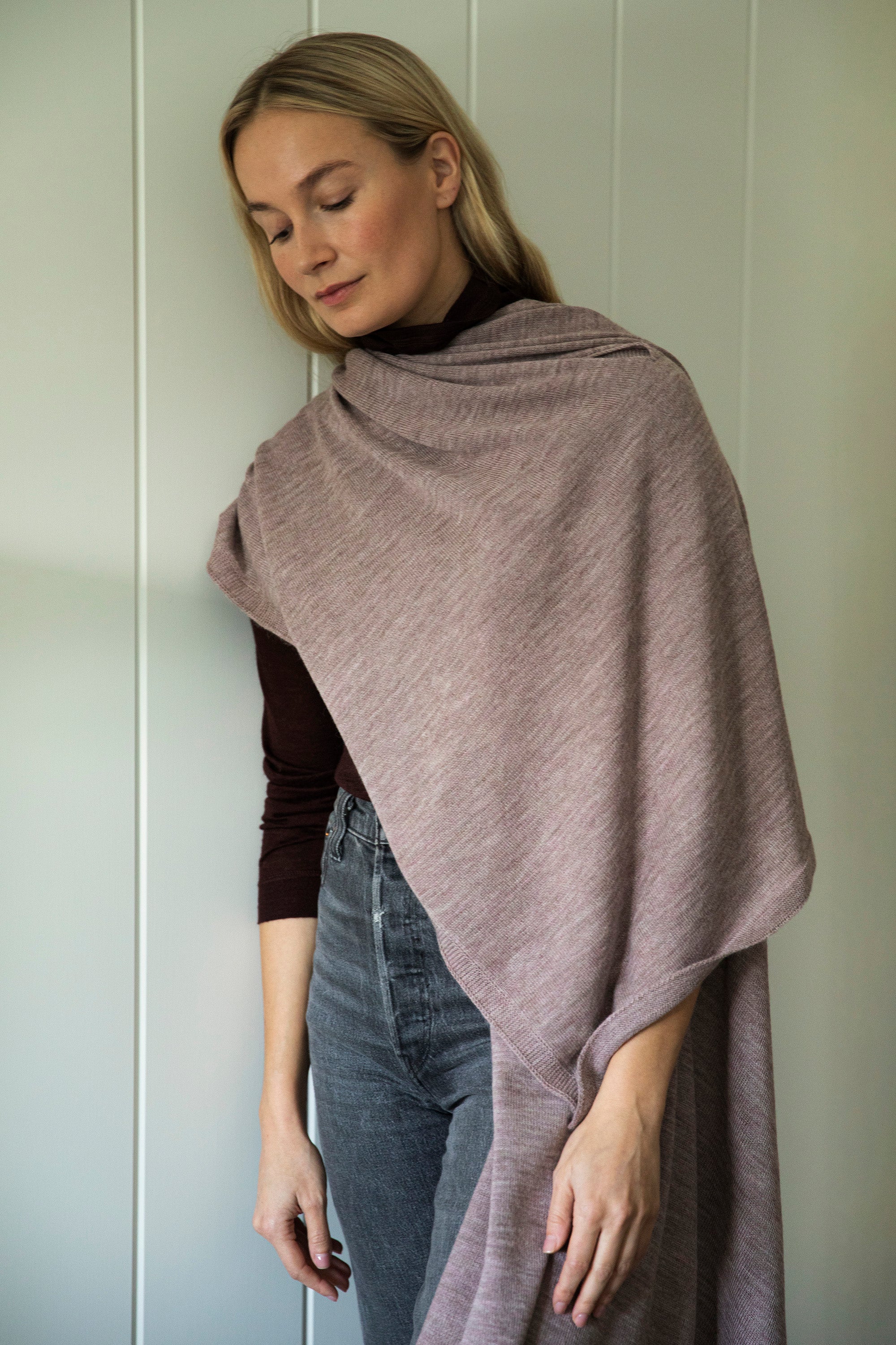 Model standing against a wall, wearing the mauve Vellie wrap draped over one shoulder. Ethically made in Peru from earth friendly fibers such as 100% baby alpaca.