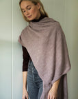 Model standing against a wall, wearing the mauve Vellie wrap draped over one shoulder. Ethically made in Peru from earth friendly fibers such as 100% baby alpaca.