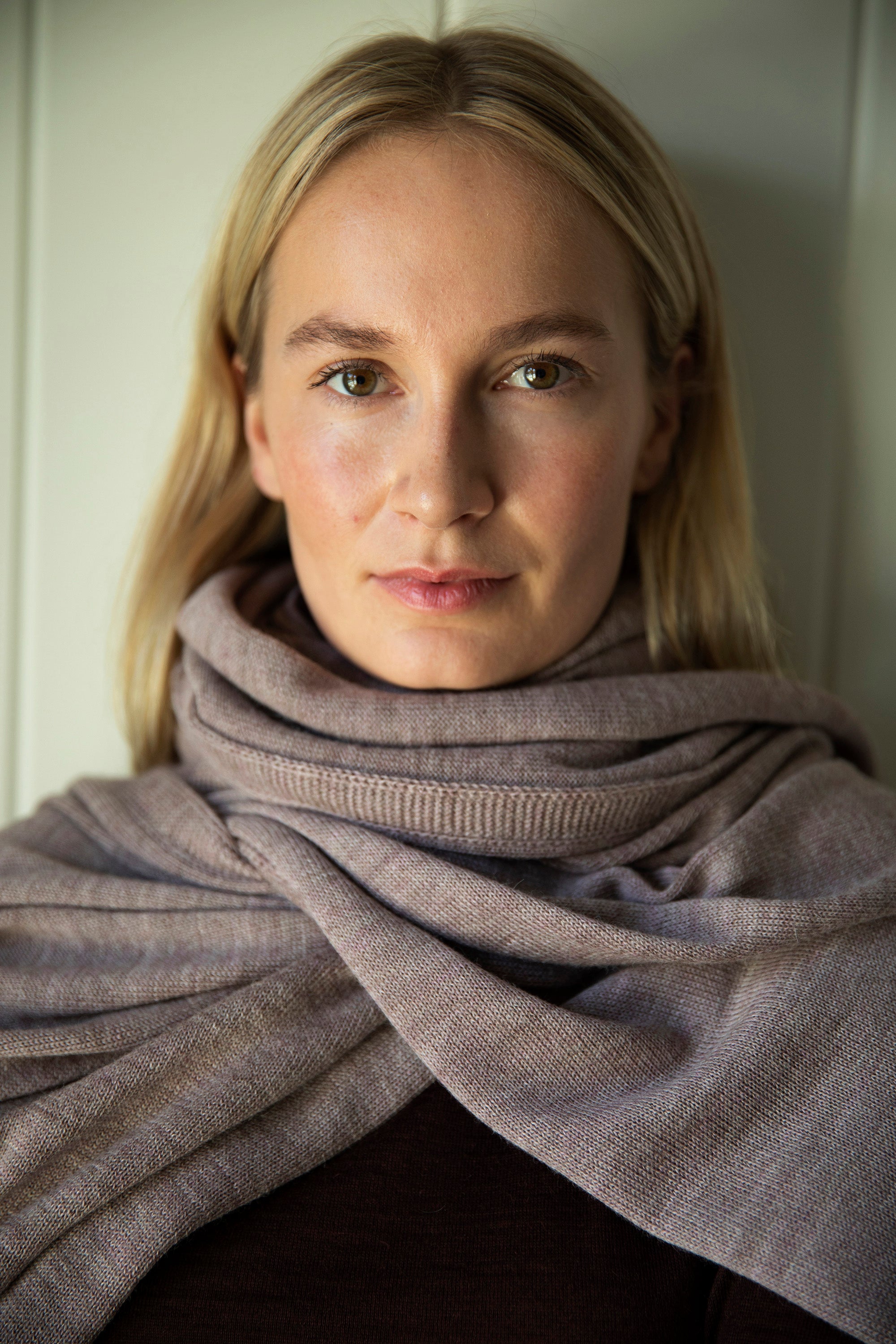 Model looking directly into the camera, wearing the mauve Vellie wrap styled as a scarf. Crafted from hypoallergenic, antimicrobial baby alpaca fiber, offering luxurious warmth.