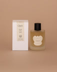 Vellie Open Spray. A wrap spray with herbal, citrus and green notes. Seen here, the transparent vessel with a black lid next to its packaged box. Box colors are neutral, cream, black and white.