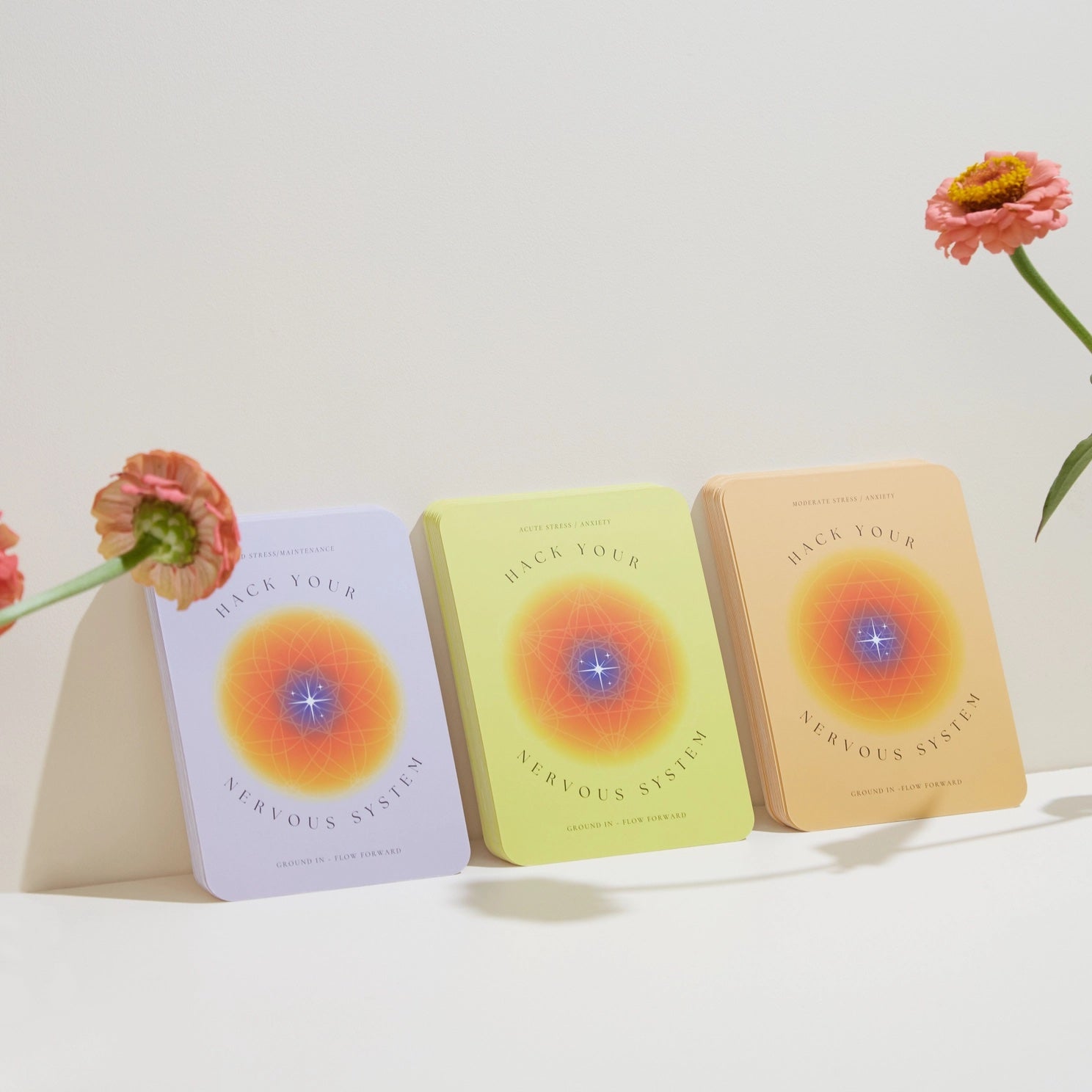 Three cards from the 'Hack Your Nervous System' deck with colorful covers on a white surface, accompanied by flowers.