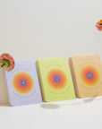 Three cards from the 'Hack Your Nervous System' deck with colorful covers on a white surface, accompanied by flowers.