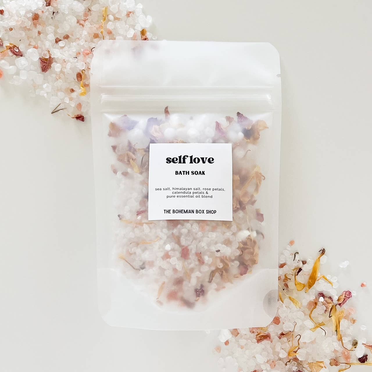 Self Love bath soak by The Bohemian Box Shop. Made in USA all natural ingredients: sea salt, Himalayan salt, rose, petals, calendula petals and pure essential oil blend. Salts are transparent color and light pink with yellow calendula petals.