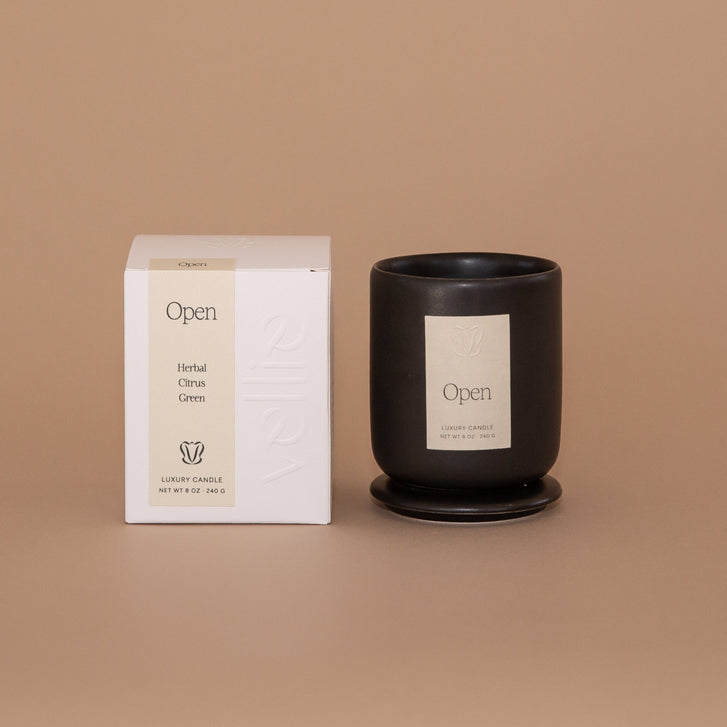 Vellie Open Candle. A luxury candle with herbal, citrus and green notes. Seen here, the chic black vessel next to its packaged box. Box colors are neutral, cream, black and white.