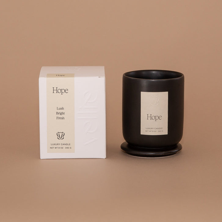 Vellie Hope Candle. A luxury candle with lush, bright and fresh notes. Seen here, the chic black vessel next to it's packaged box. Box colors are neutral, cream, black and white.