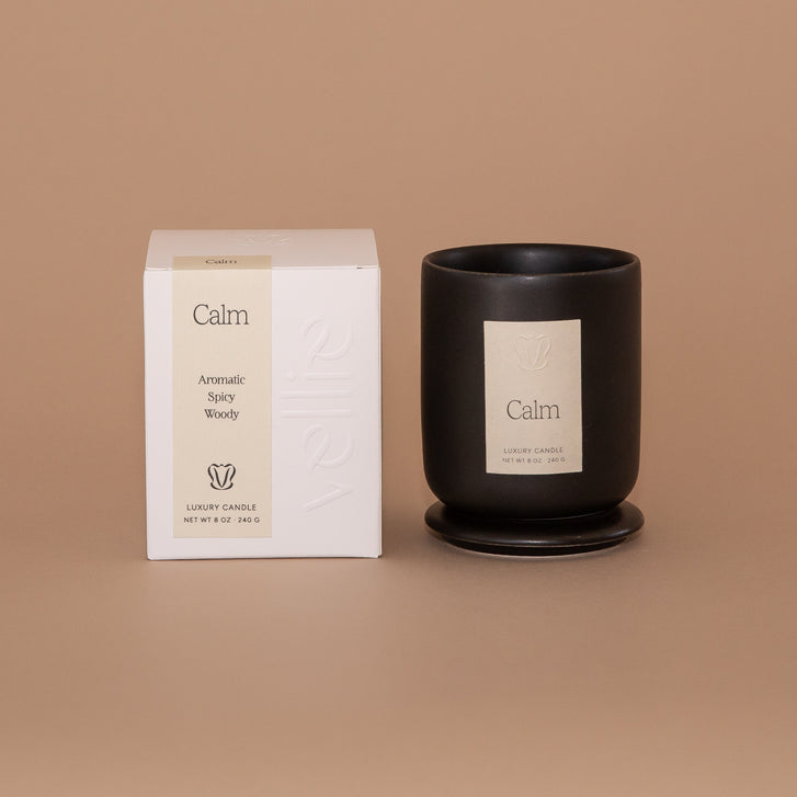 Vellie Calm Candle. A luxury candle with aromatic, spicy and woody notes. Seen here, the chic black vessel next to it's packaged box. Box colors are neutral, cream, black and white.