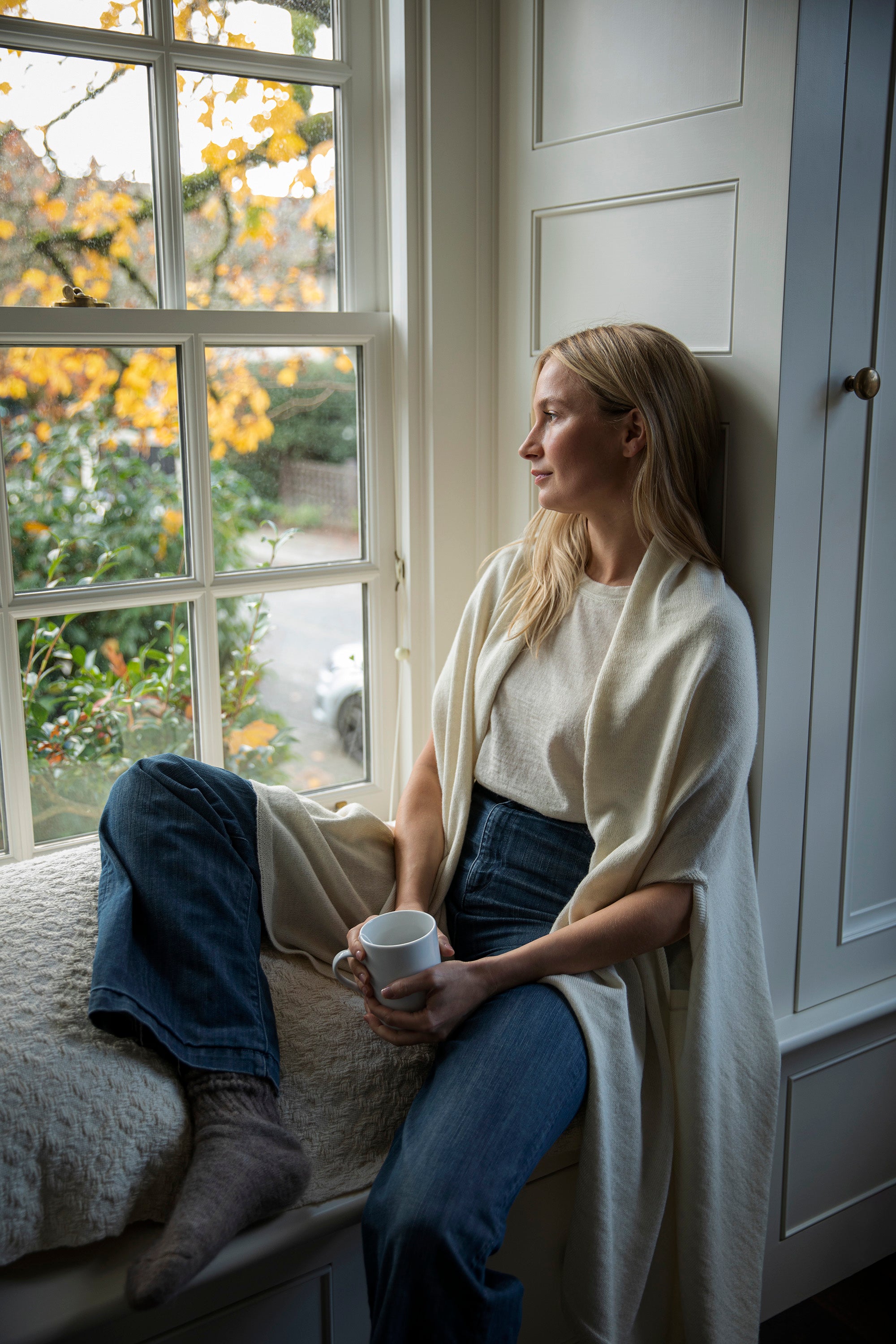 Model holds a cup of tea as she sits inside a window nook looking outside the window. She wears a long ivory wrap that comforts her. Crafted from 100% fair trade, sustainably sourced baby alpaca.