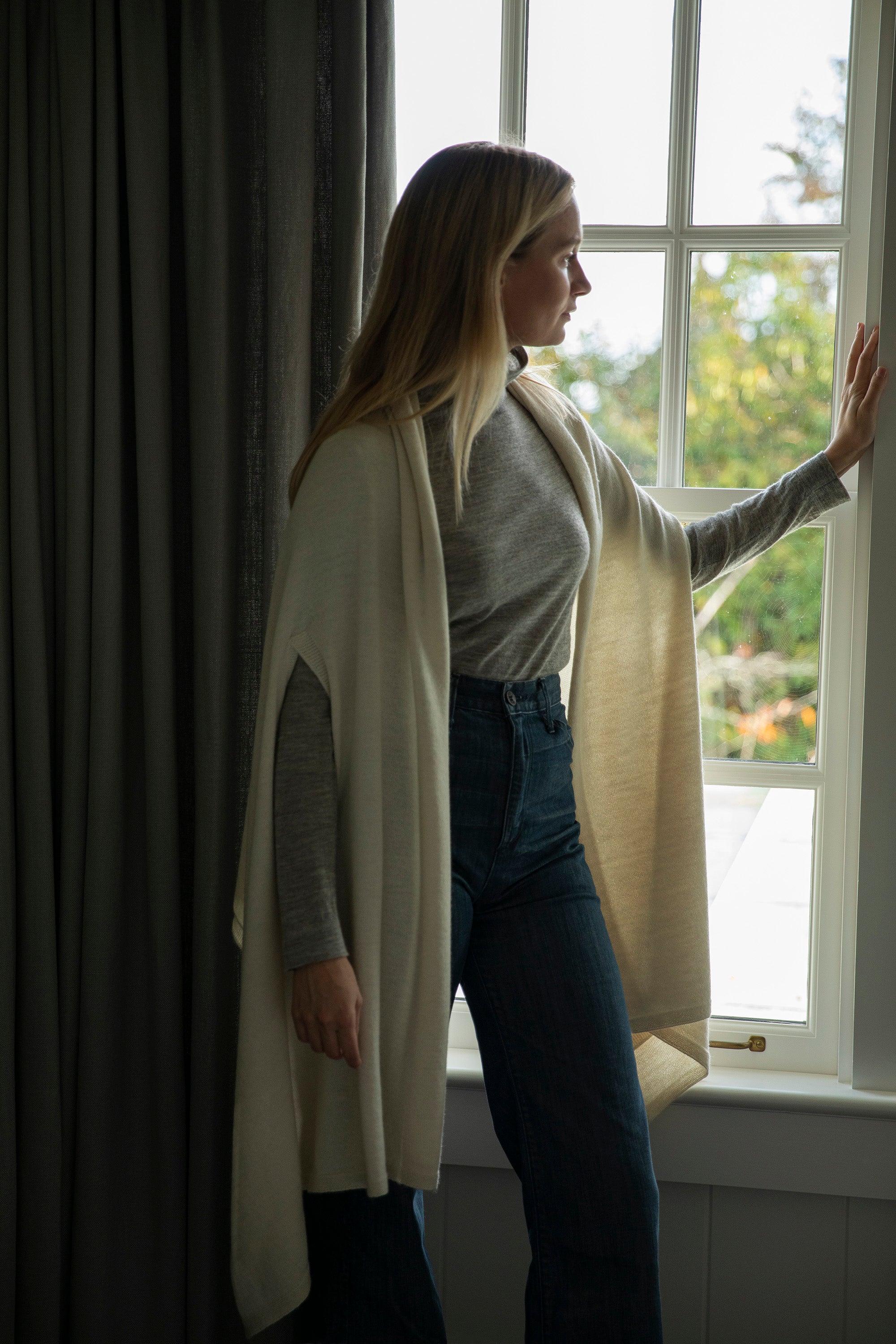 Model wearing Vellie ivory wrap, gently pulling back blue curtains and gazing pensively out the window, embodying the comfort and warmth of quiet sustainable luxury.