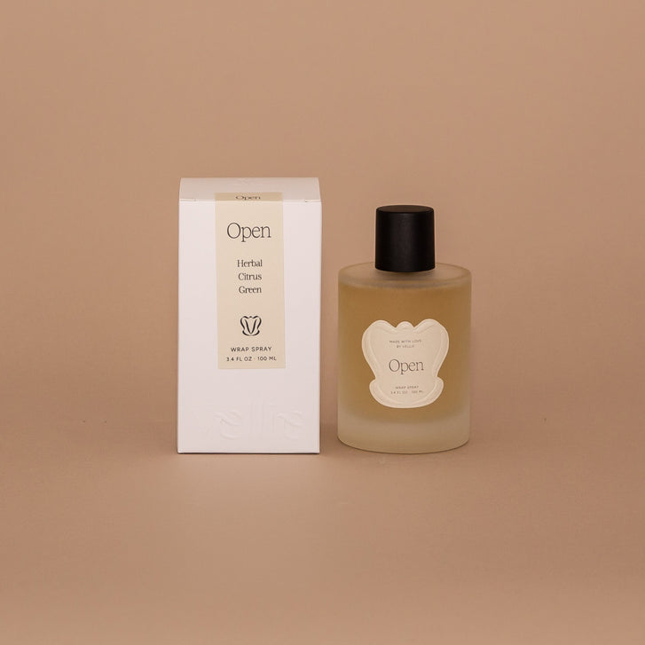 Vellie Open Spray. A wrap spray with herbal, citrus and green notes. Seen here, the transparent vessel with a black lid next to its packaged box. Box colors are neutral, cream, black and white.