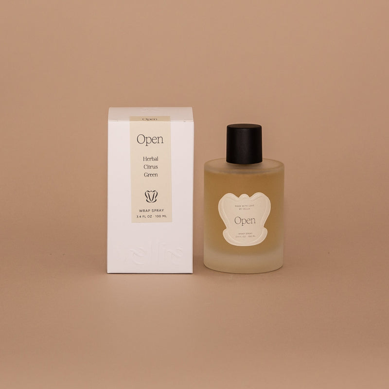 Vellie Open Spray. A wrap spray with herbal, citrus and green notes. Seen here, the transparent vessel with a black lid next to its packaged box. Box colors are neutral, cream, black and white.