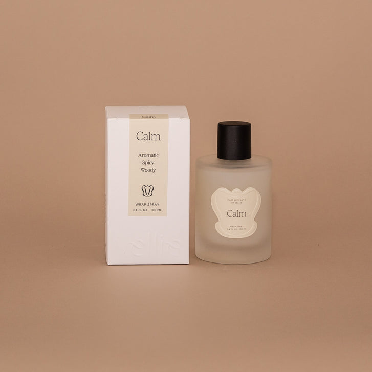 Vellie Wrap Spray in Calm scent next to the packaged box. The spray is showcased  in a 3.4 oz bottle, vegan and cruelty-free. It contains a grounding blend of bergamot, lavender, and sandalwood, perfect for enhancing the embrace of your wrap.