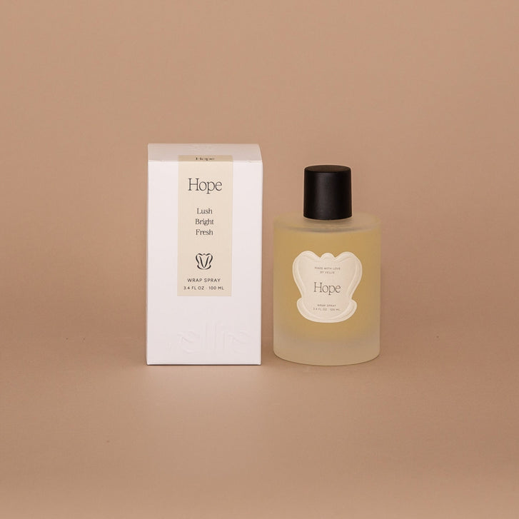 Vellie Wrap Spray in Hope next to neutral pacakged box. The spray is showcased  in a 3.4 oz bottle, vegan and cruelty-free. It contains a grounding blend of bergamot, lemon and ylang ylang perfect for enhancing the embrace of your wrap.